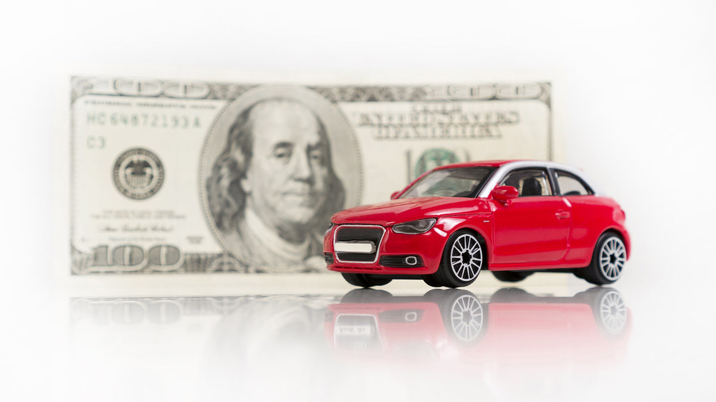 Red toy car in front of a $100 bill – Exploring the impact of rising car insurance rates in 2025.
